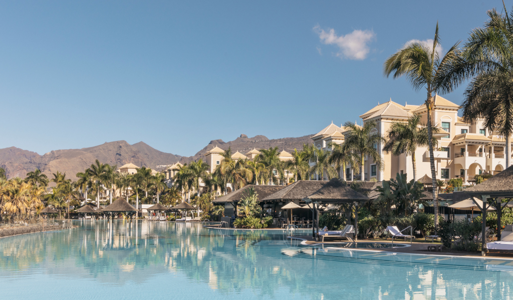 Piscina y palmeras frente a un hotel en Tenerife, con montaña al fondo.