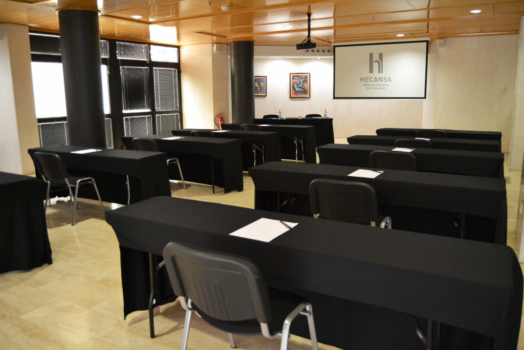 Sala de conferencias con mesas negras, sillas y pantalla de proyección en Hecansa, Tenerife.