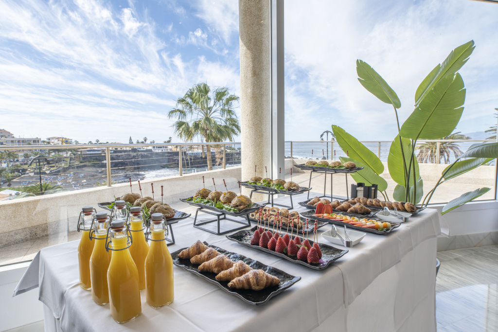 Mesa con desayuno buffet frente al mar en Tenerife, con jugos y pasteles variados.