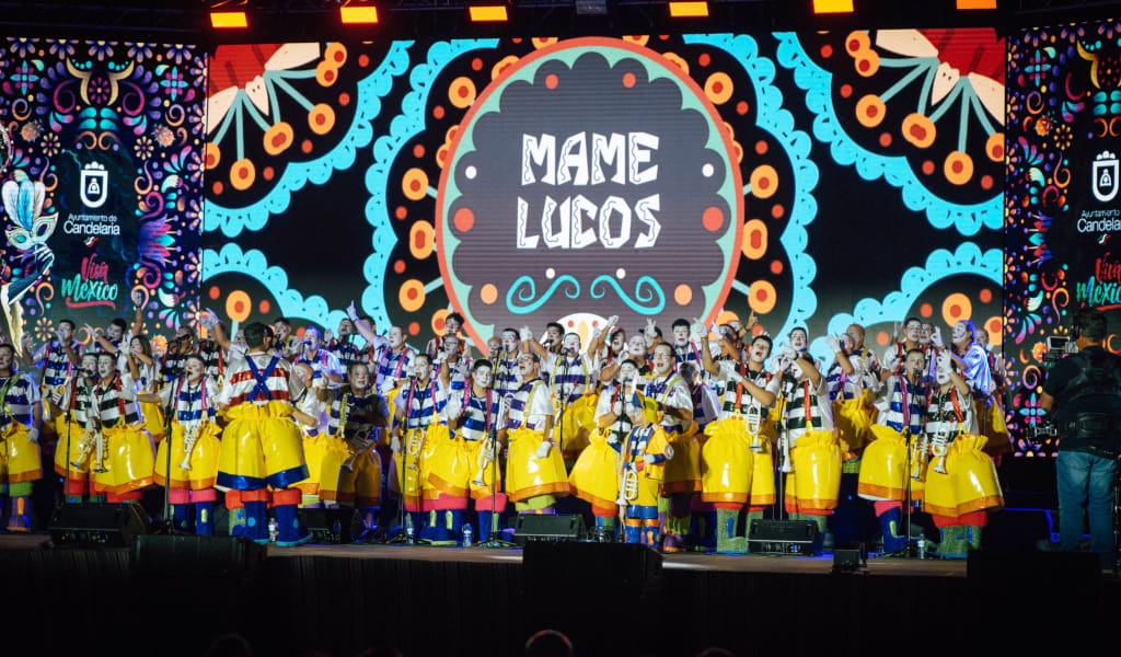 Grupo de personas en el escenario de Candelaria, con fondo colorido y texto Mame Luicos.