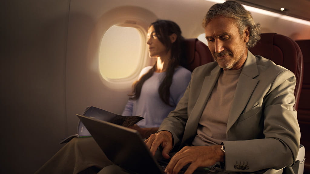 Hombre usando laptop junto a mujer leyendo en un avión con luz natural.