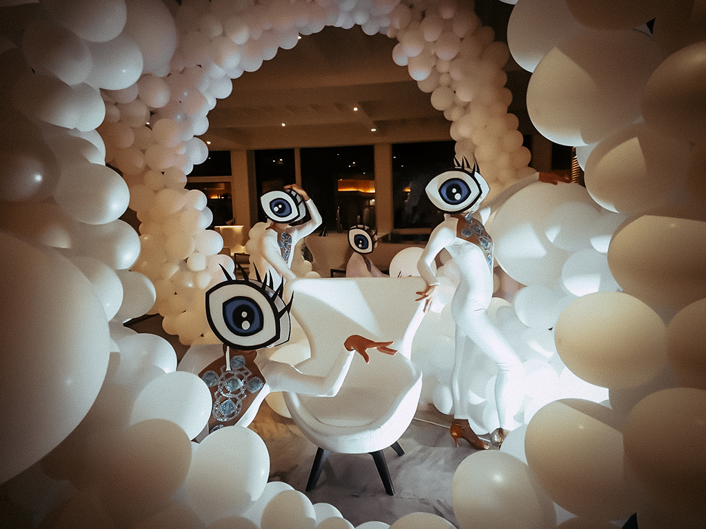 Personas con disfraces de ojos gigantes rodeadas de globos blancos en un salón.