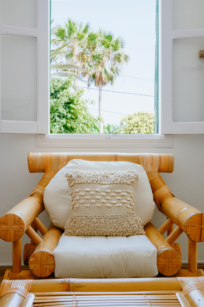 Sillón de mimbre con cojín en sala frente a ventana abierta con vista a palmera en Tenerife.