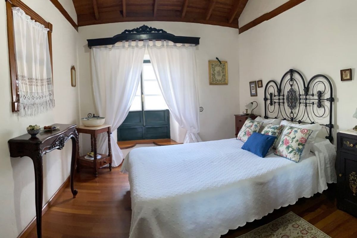 Habitación con cama de hierro forjado, cojines florales y ventana con cortinas blancas en Tenerife.