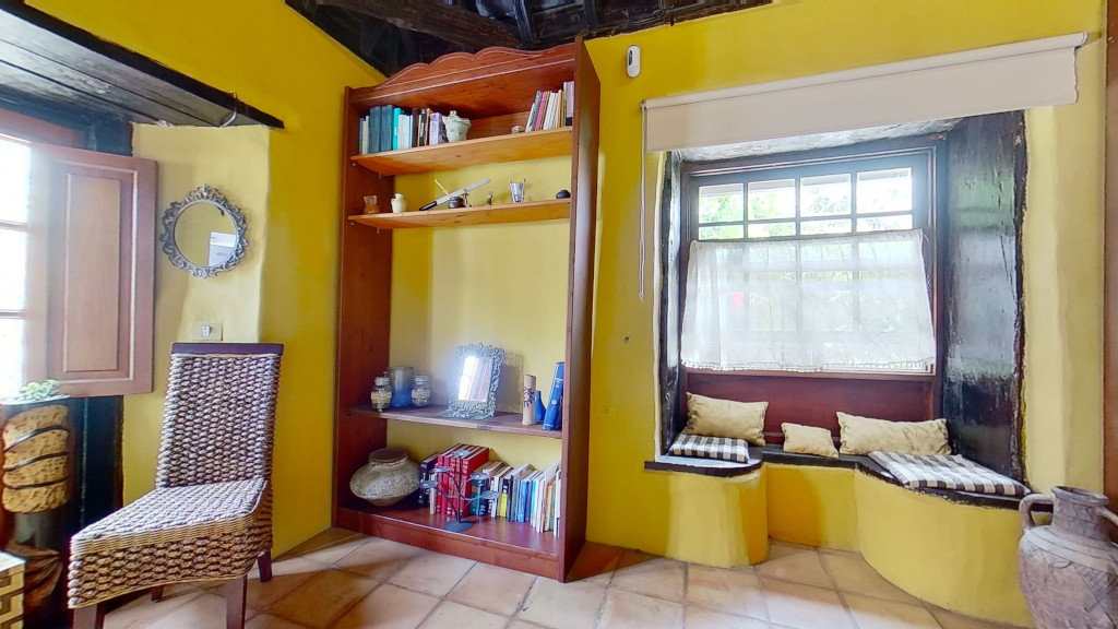 Sala amarilla en Tenerife con estantería, silla tejida y ventana con cojines a cuadros.