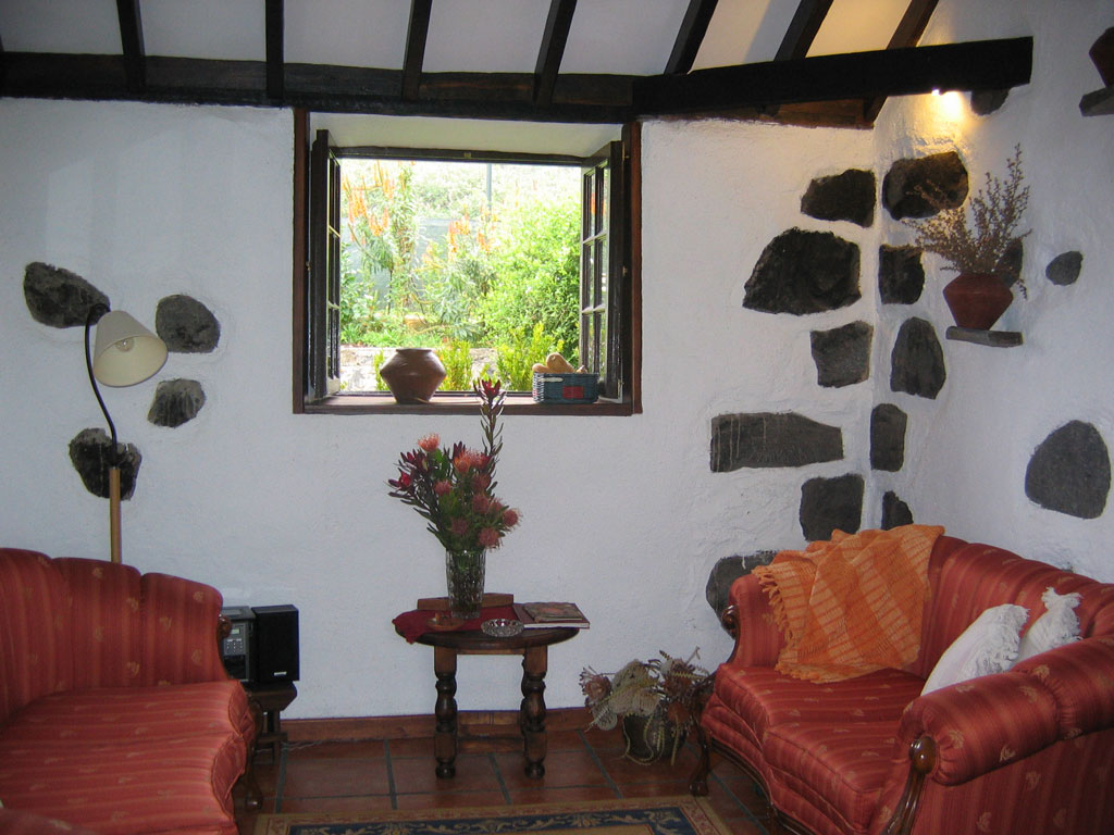Sala de estar rústica en Tenerife con sofá rojo, ventana y plantas exteriores.