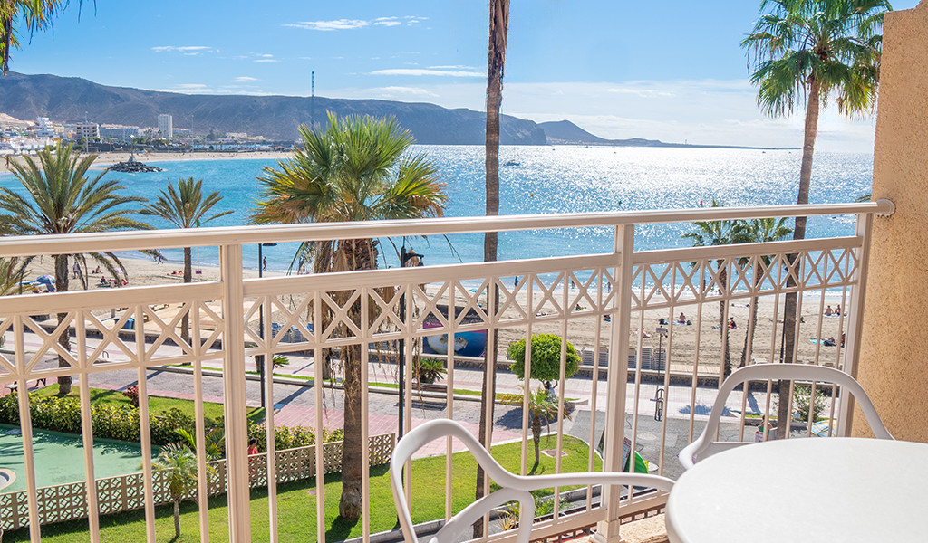 Vista desde un balcón hacia la playa de Los Cristianos en Tenerife, con mar y palmeras.