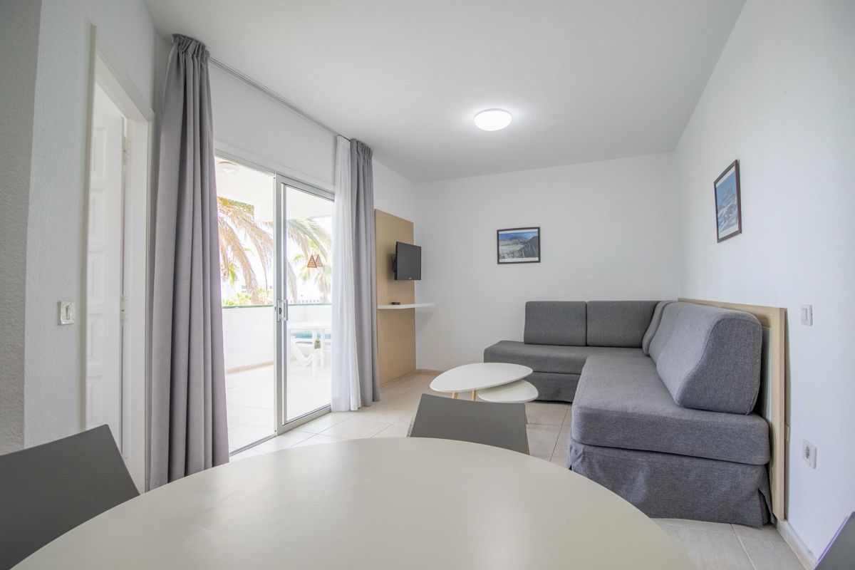 Sala de estar con sofá gris y mesa blanca, puerta a terraza con palmeras en Tenerife.
