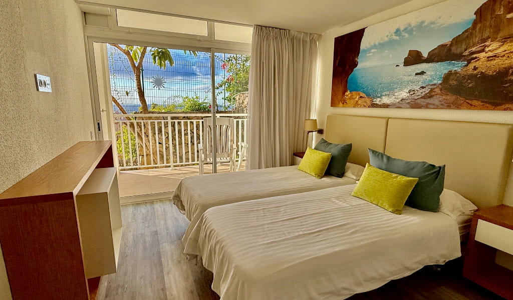Habitación de hotel con camas individuales, balcón y cuadro de paisaje costero en Tenerife.
