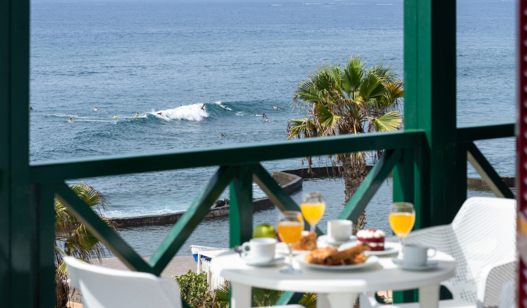 Desayuno en terraza con vista al mar en Tenerife, palmera y surfistas de fondo.