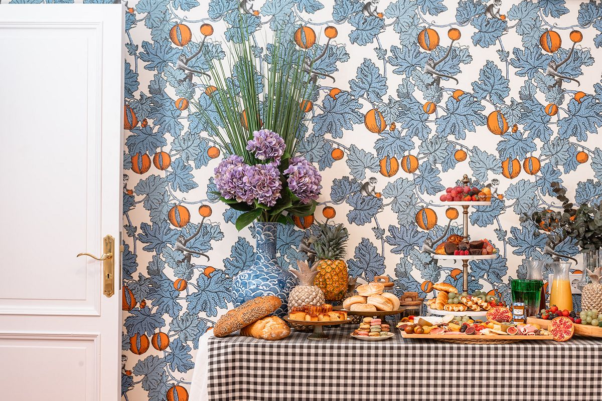 Mesa con platos variados de frutas, panes y dulces frente a papel tapiz decorado.