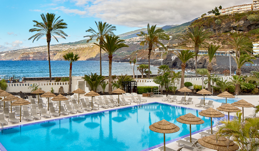Piscina al aire libre con vista al mar en Tenerife, rodeada de tumbonas y palmeras.
