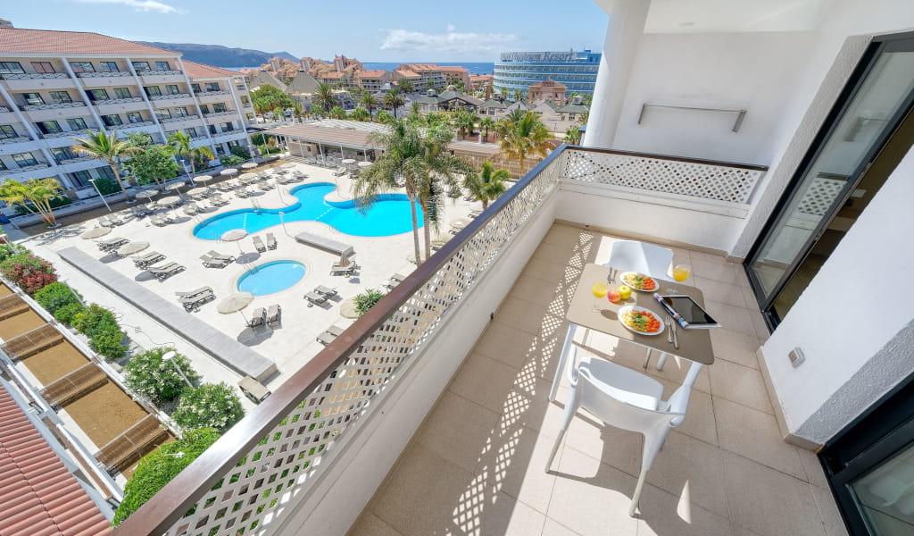 Balcón con vista a la piscina del hotel en Tenerife, mesa con platos y bebidas.
