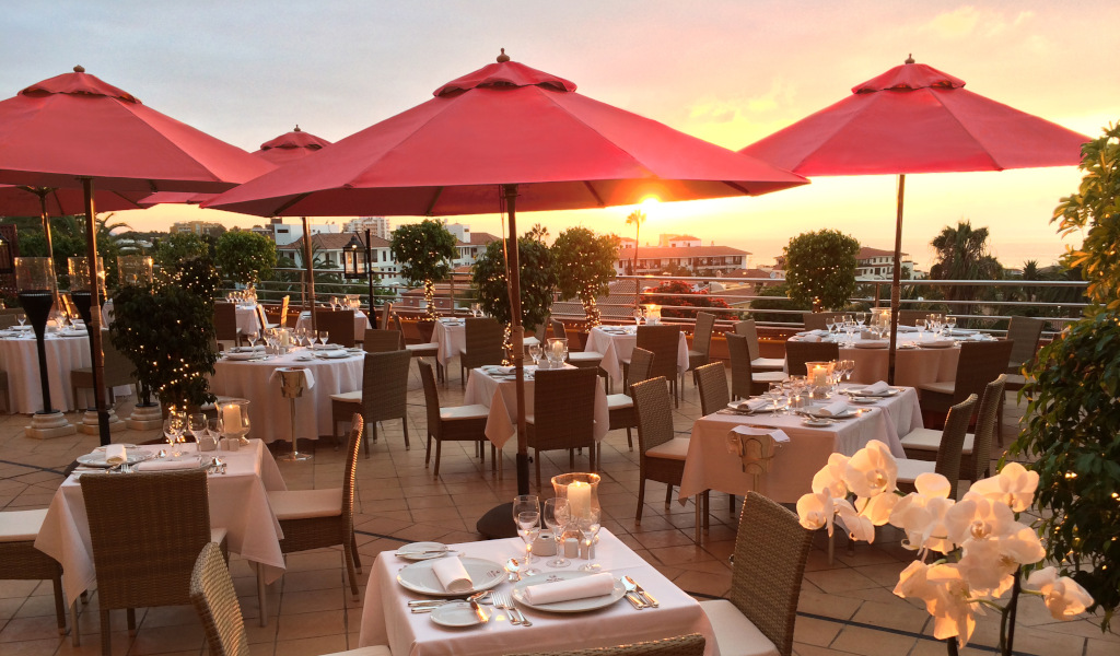 Terraza de restaurante en Tenerife al atardecer con mesas y sombrillas rojas.