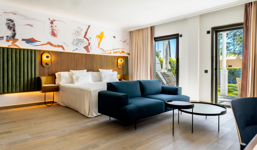 Habitación moderna en Tenerife con cama doble, sofá, mesas y balcón con vista al jardín.