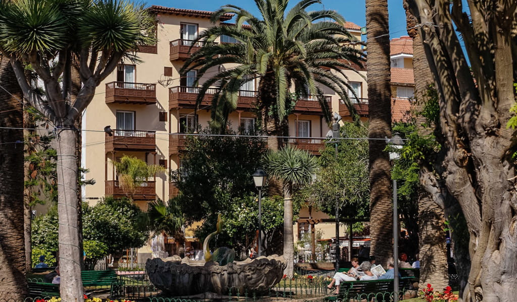 Plaza arbolada con edificio y personas descansando en Puerto de la Cruz, Tenerife.