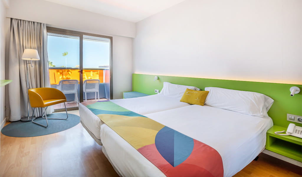 Habitación de hotel en Tenerife con dos camas, sillón amarillo y balcón.