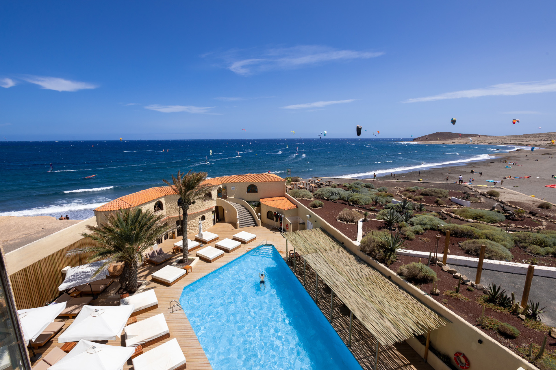 Vista de la playa El Médano en Tenerife con kitesurfistas, piscina y terraza cercana.