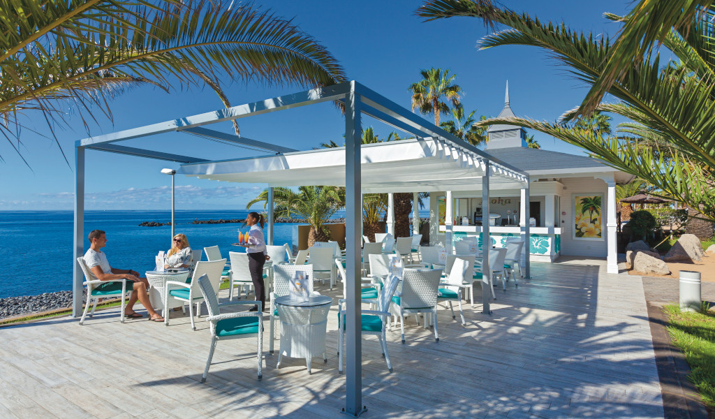 Terraza con mesas y sillas junto al mar en Tenerife, servidor lleva bebidas.