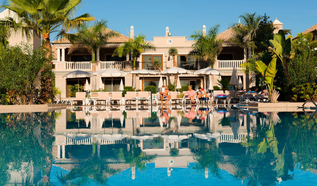 Piscina con tumbonas y sombrillas frente a hotel rodeado de palmeras en Tenerife.