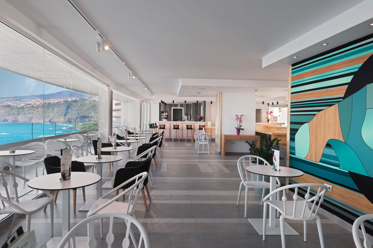 Interior de cafetería moderna con vista al mar en Tenerife, mesas y sillas blancas.