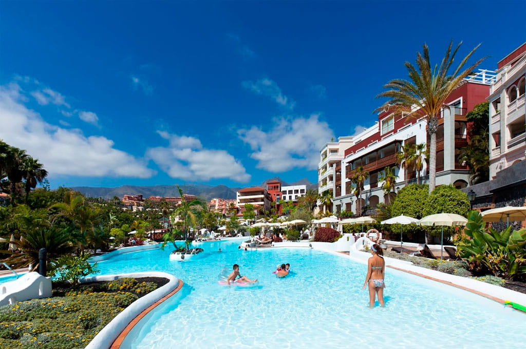 Piscina rodeada de palmeras y edificios en un hotel de Tenerife.