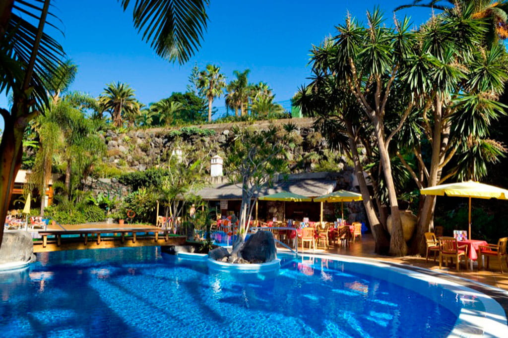 Piscina rodeada de palmeras y mesas con sombrillas en un resort en Tenerife.