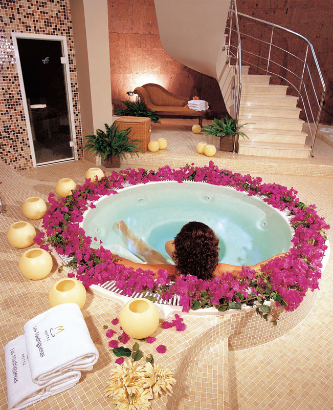 Persona relajándose en un jacuzzi rodeado de flores, toallas etiquetadas Les Meridien.