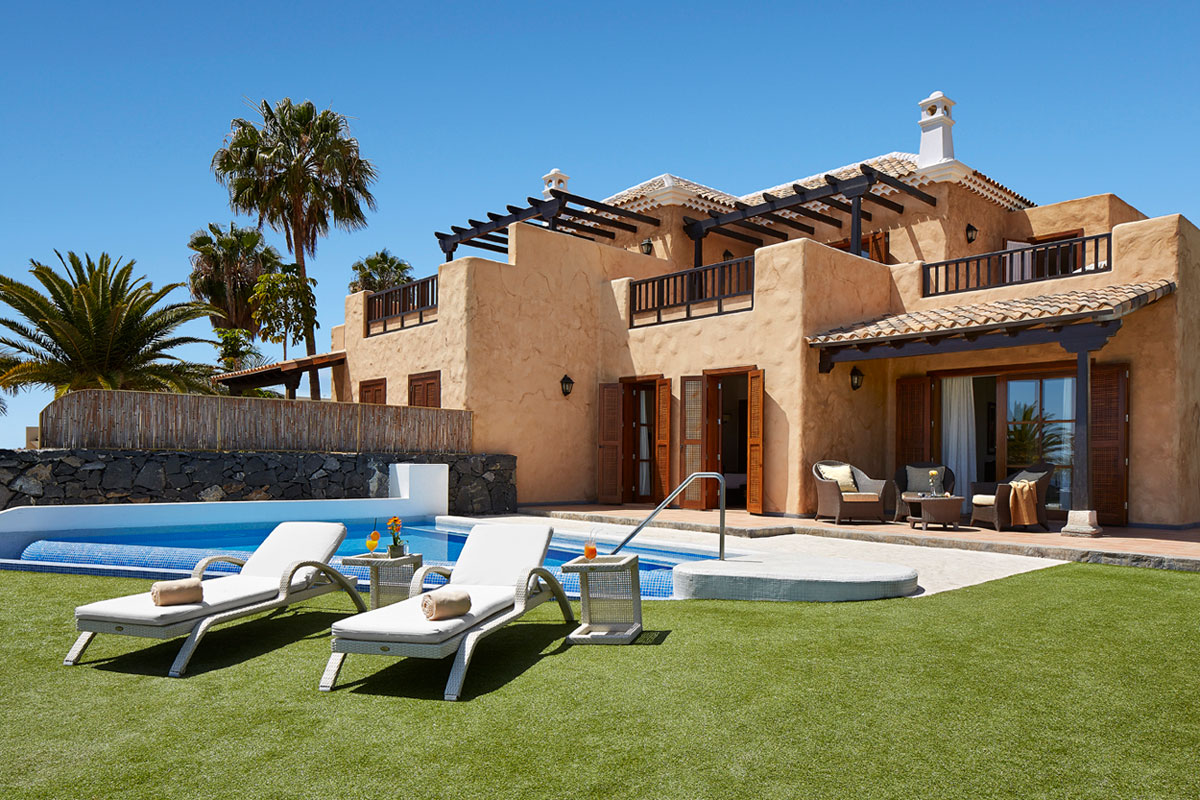 Villa con piscina en Tenerife, césped y tumbonas al frente, rodeada de palmeras.