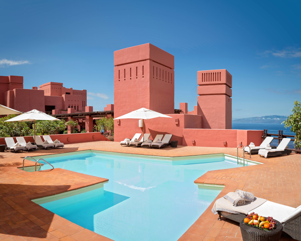 Piscina rodeada de tumbonas y edificios rojo terracota con vista al mar en Tenerife.