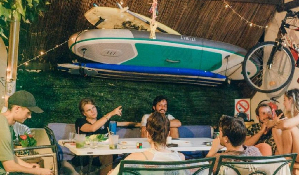 Grupo de personas charlando en una terraza decorada, con tabla de surf y bicicleta colgadas.
