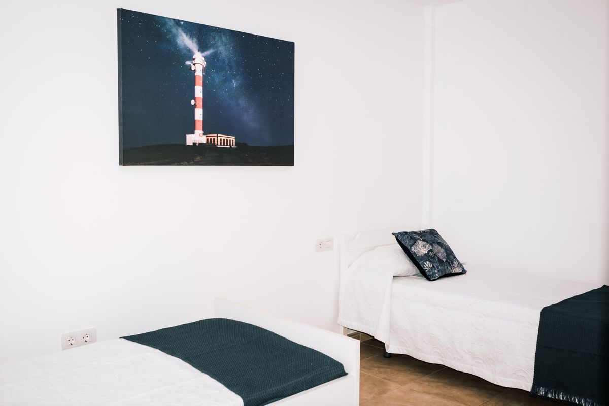 Cama doble con colcha blanca y cuadro de un faro en habitación de Tenerife.