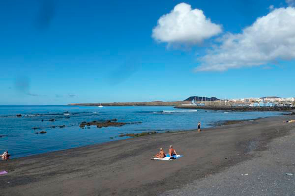 Playa con personas descansando en Los Cristianos, Tenerife, bajo un cielo azul.