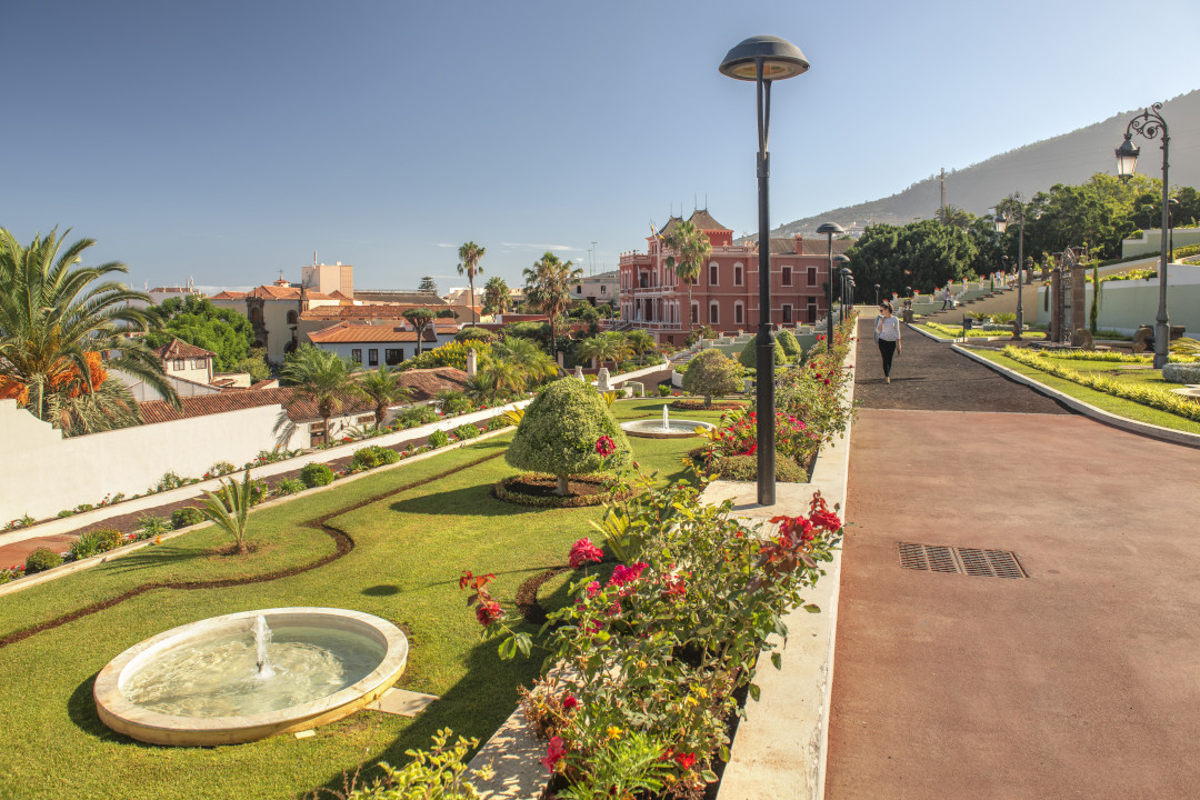 Jardines y edificio rojo en Puerto de la Cruz, Tenerife, con montañas al fondo.