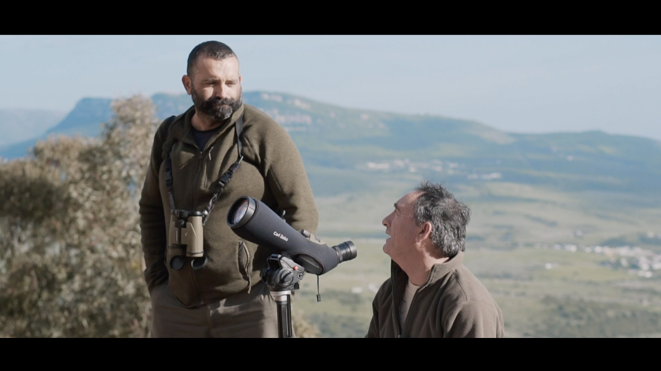 Dos hombres con binoculares y telescopio en paisaje montañoso.
