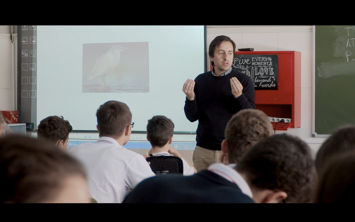 Profesor dando clase frente a estudiantes con imagen de pájaro en pantalla.
