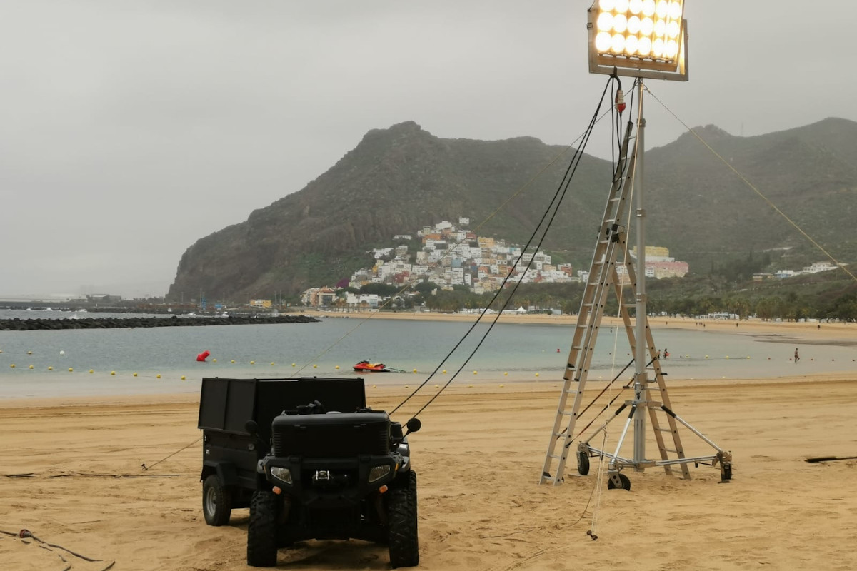 Luces y vehículo en la playa de Las Teresitas, Tenerife, con casas y montaña al fondo.