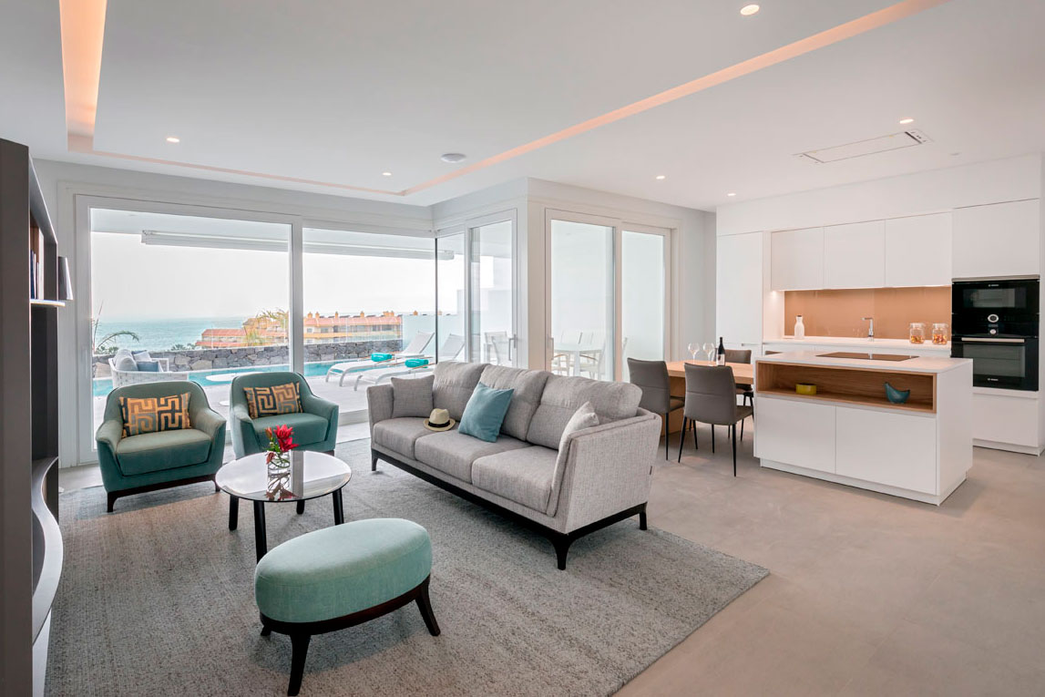 Sala de estar moderna con vista al mar en Tenerife, sofás grises y cocina integrada.