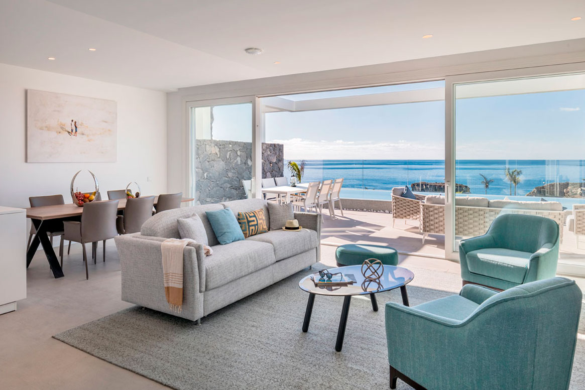 Sala de estar moderna con vista al mar en Tenerife. Sofá gris, sillas azules y mesa.
