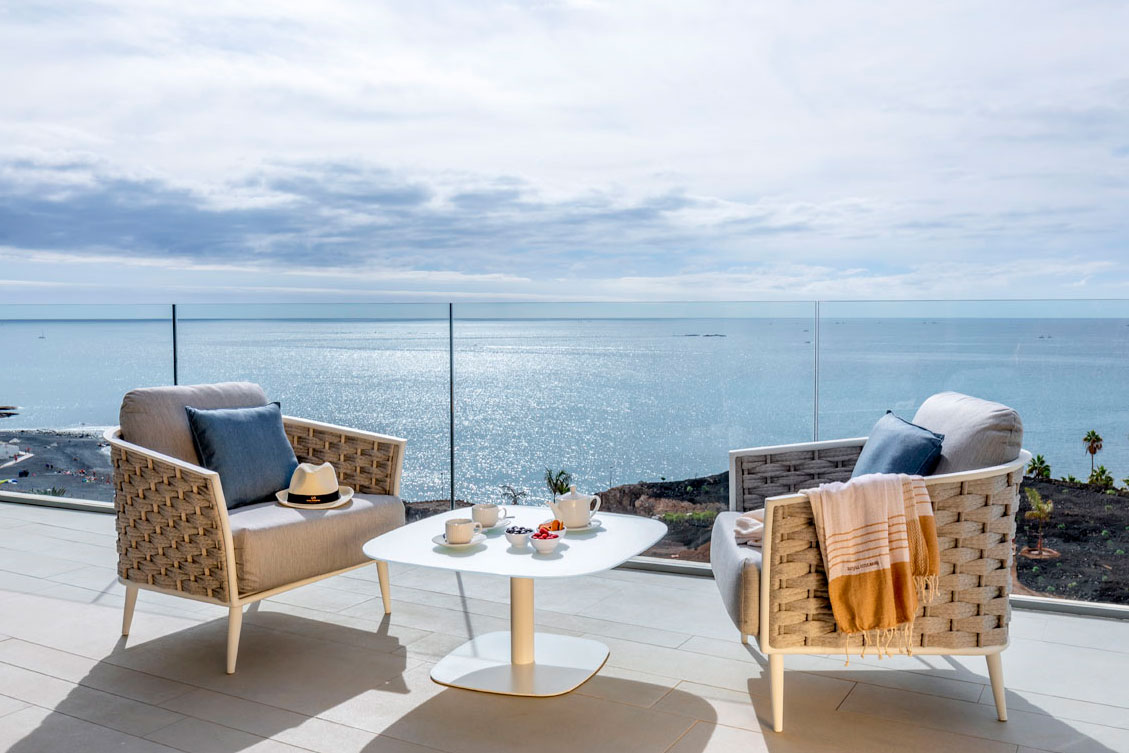 Terraza con sillas y mesa frente al mar en Tenerife, fondo azul y cielo nublado.