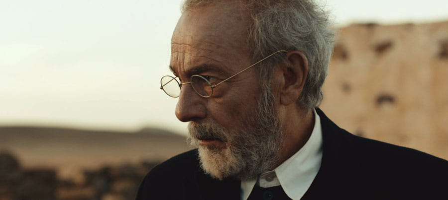 Un hombre con gafas y barba mirando hacia la derecha en un entorno árido.