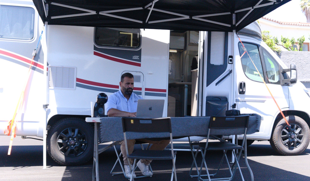 Persona trabajando en computadora portátil bajo toldo junto a caravana en Tenerife.