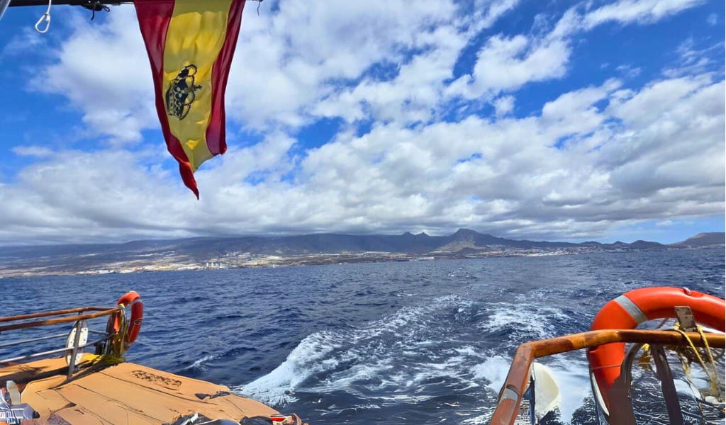 Velero en el mar con bandera de España y vista de la costa de Tenerife bajo cielo nublado.