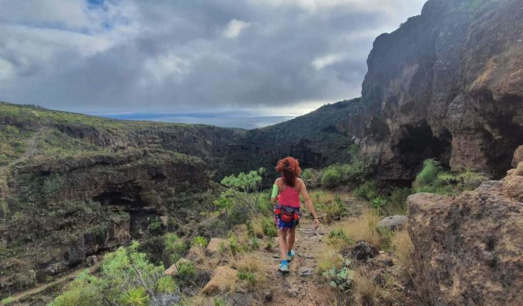 Persona caminando por un sendero rocoso en un barranco de Tenerife bajo un cielo nublado.