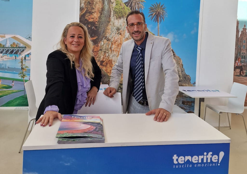 Dos personas sonriendo en un stand promocional de Tenerife con folletos en la mesa.