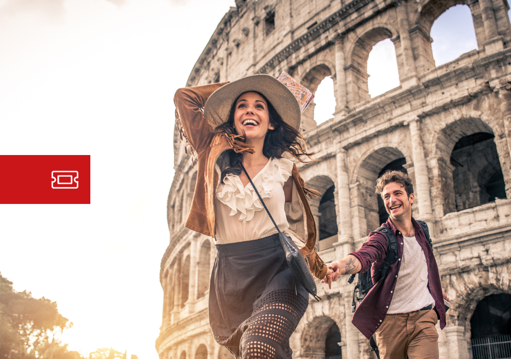 Pareja de turistas sonrientes corriendo frente al Coliseo de Roma al atardecer, con mochilas y ropa informal.