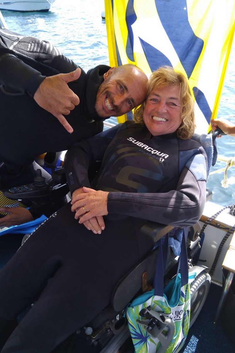 Personas en trajes de buceo sonrientes en una silla de ruedas cerca del mar en Tenerife.
