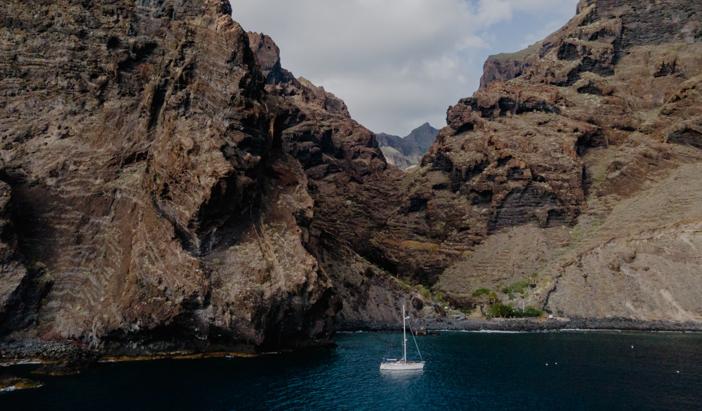 Velero en la costa rocosa de Tenerife con aguas azules y montañas al fondo.