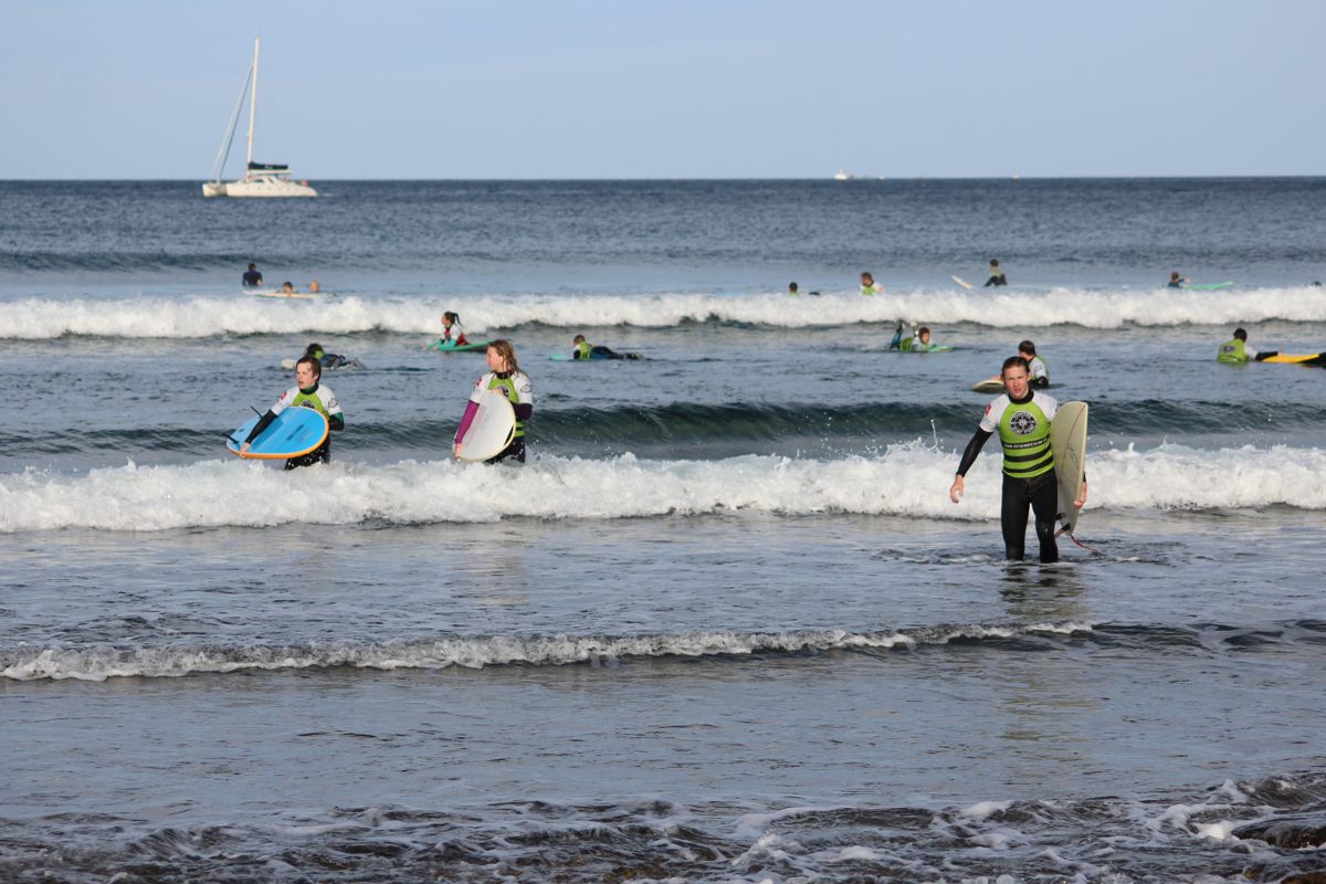 Surfistas con tablas en Playa de Las Américas, Tenerife, con velero al fondo.