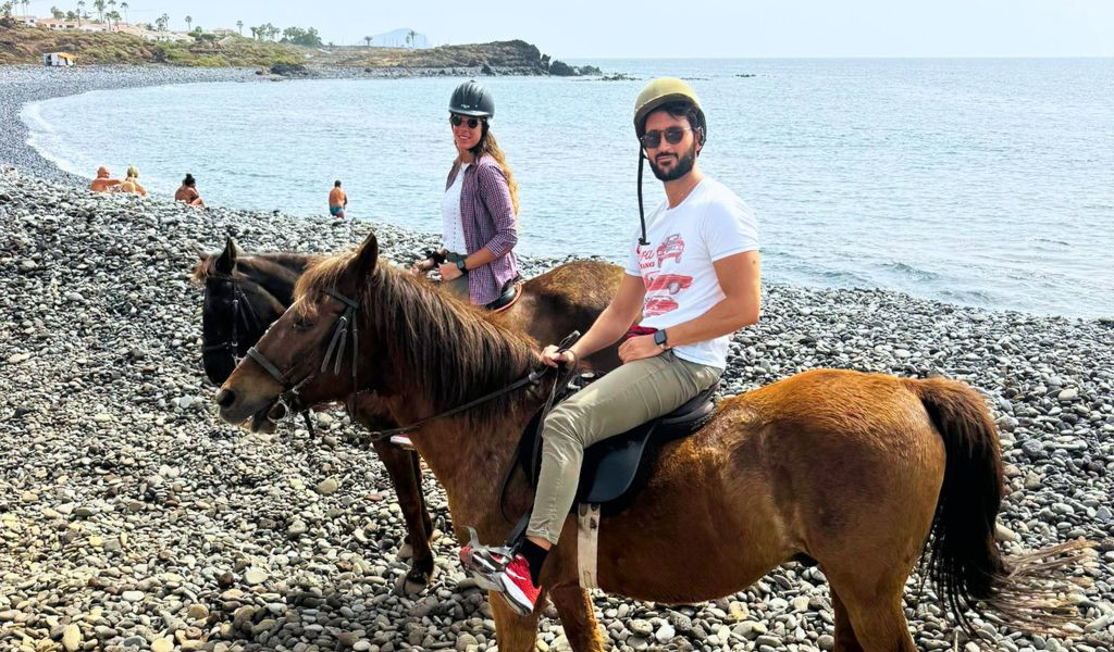 Personas montando a caballo en la playa de piedras de Tenerife, con el mar de fondo.